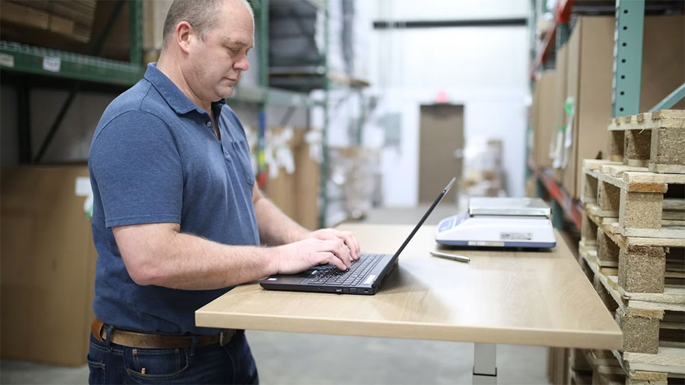 A warehouse staff member working through freight records on a laptop at a standing desk beside a parcel scale