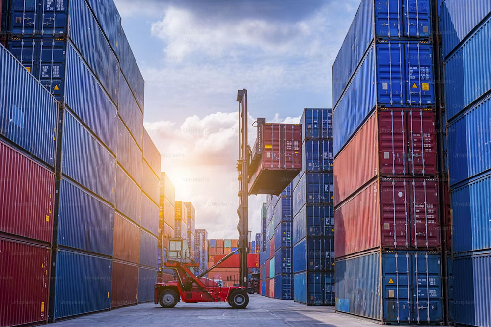 A reach stacker lifting a shipping container between stacked container rows in a freight yard