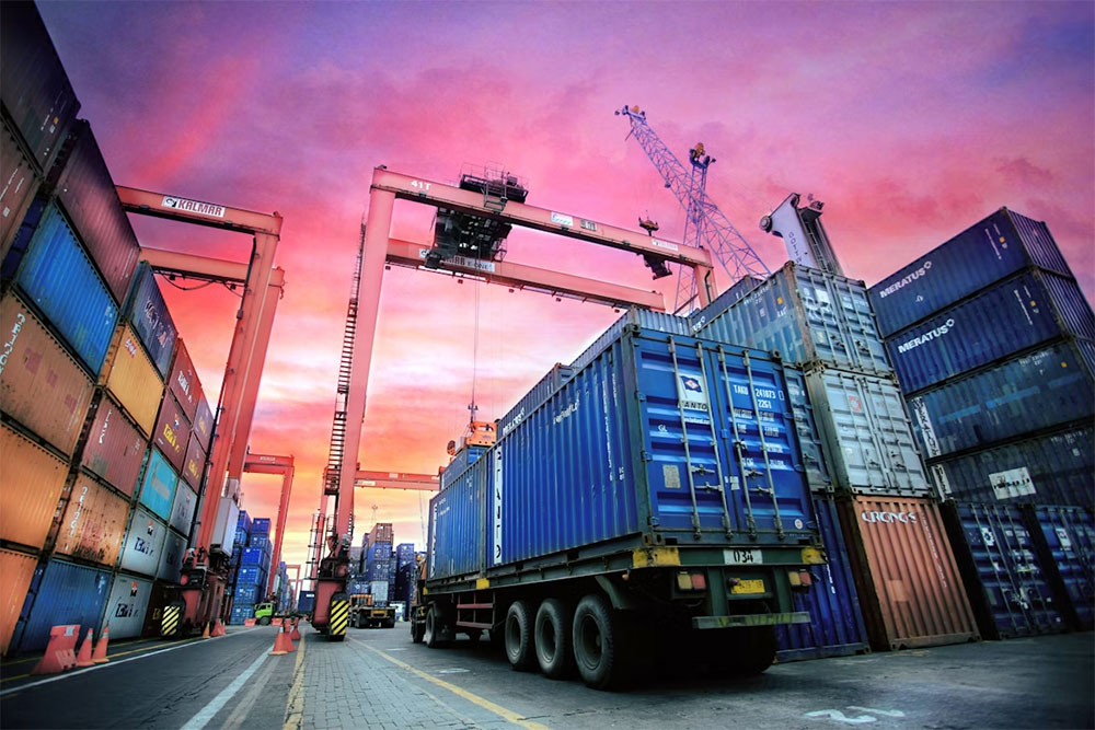A truck hauling a shipping container beneath gantry cranes in a large container terminal at sunset